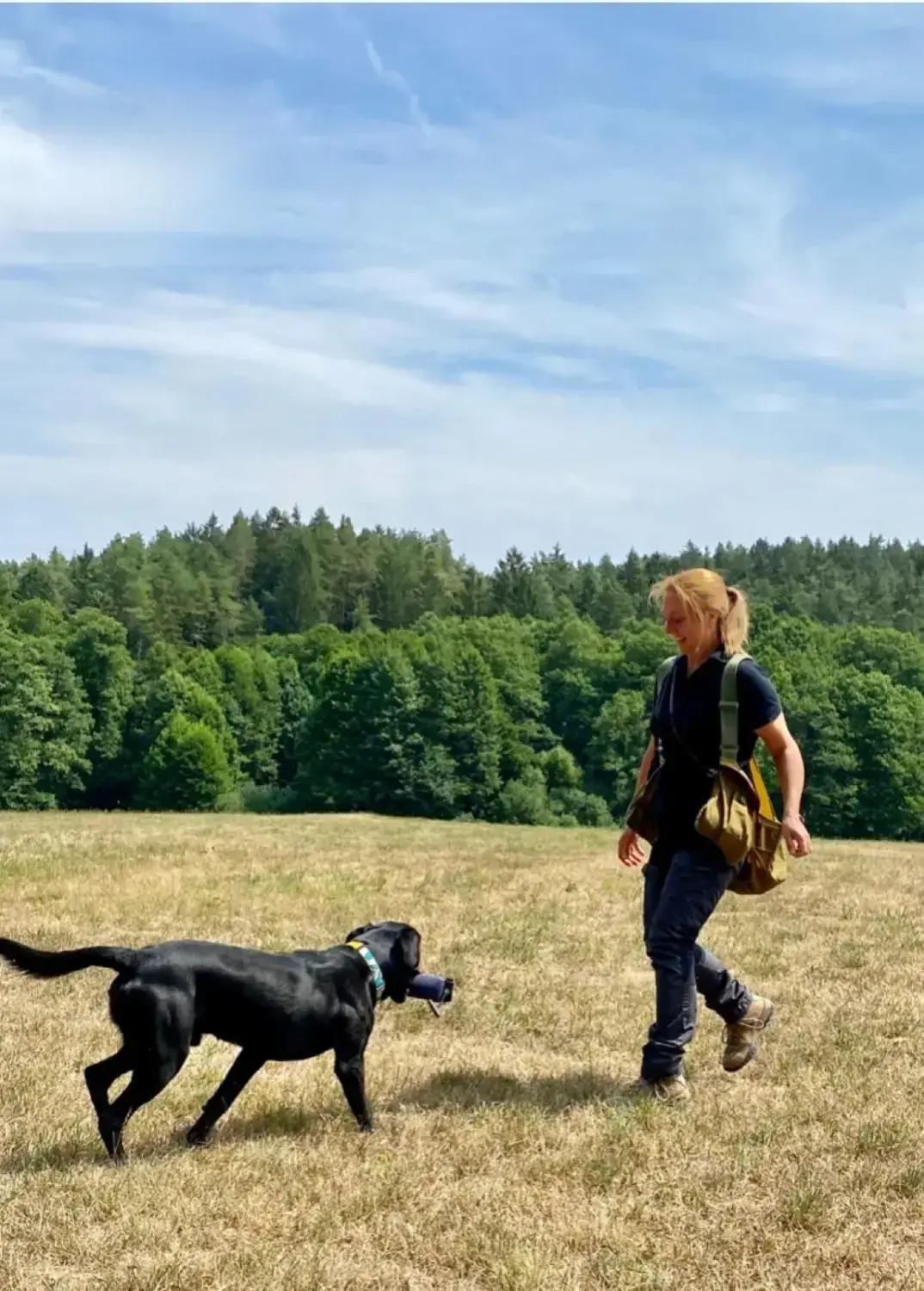 Nadine Roth geht mit ihrem schwarzen Labrador über eine sommerliche Wiese am Waldrand